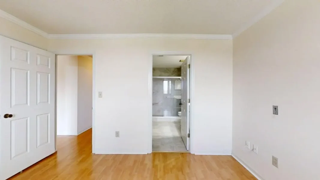 Empty bedroom with light wood flooring, left closet door, and a bathroom doorway straight ahead.