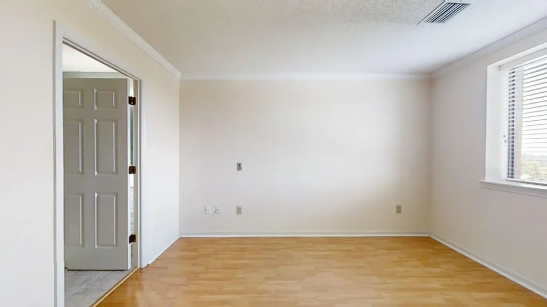 Empty bright room with light wood flooring, white walls, a window with blinds on the right, and an open doorway on the left.