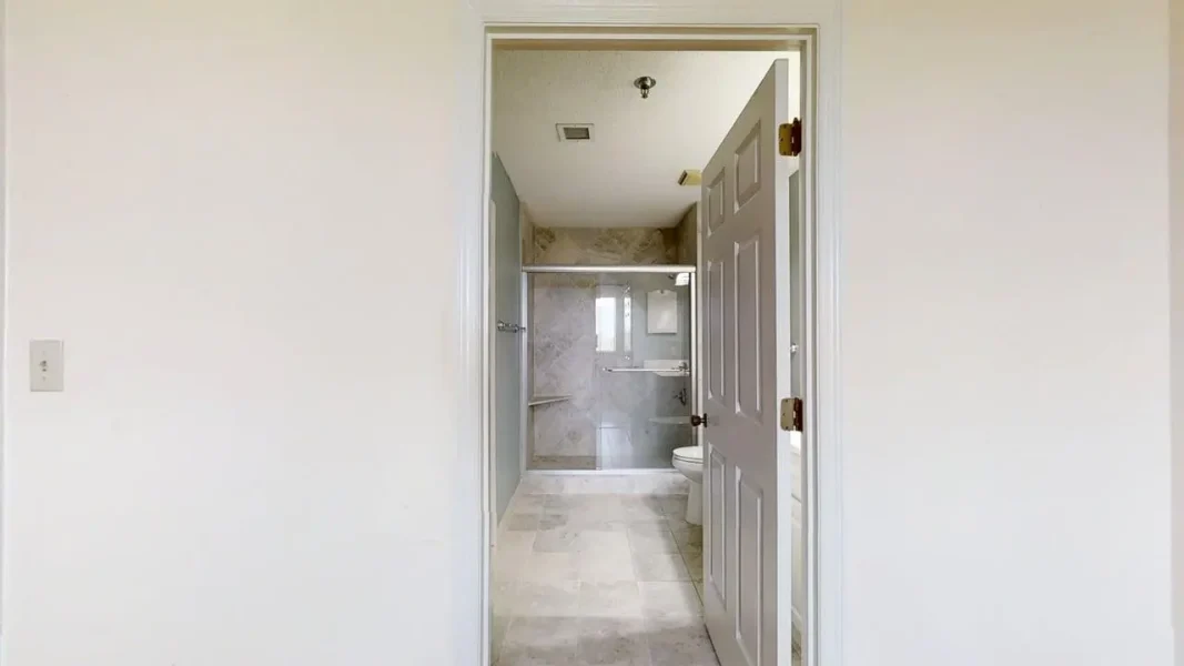 Open doorway into a modern bathroom with a glass shower enclosure, marble tiles, and a toilet visible through the doorway, neutral tones.