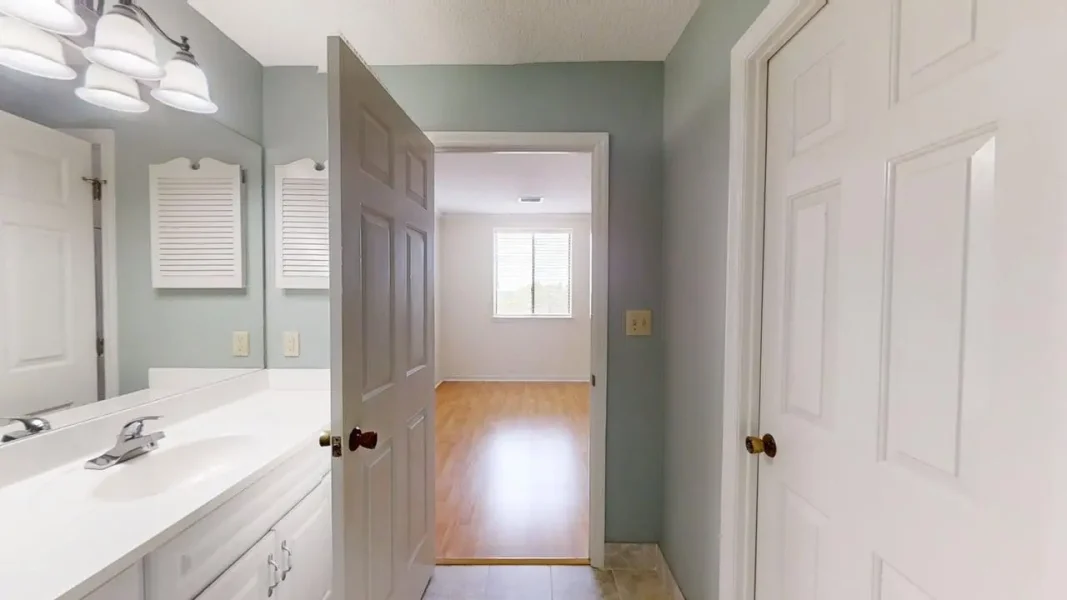 Bathroom with white double-sink vanity, mirror, and a door opening to a sunlit room with hardwood floor and a window