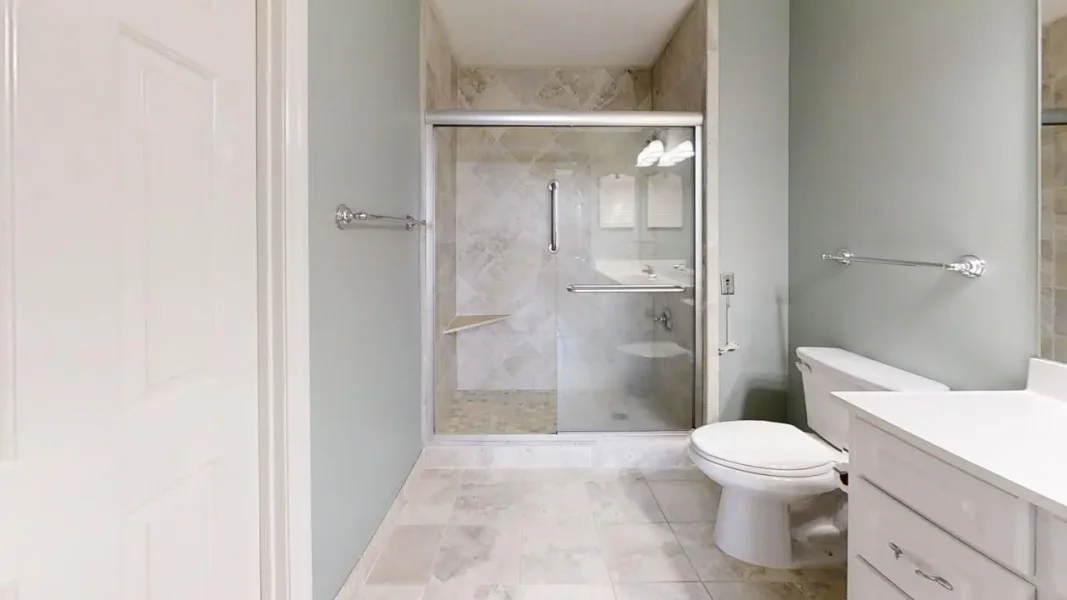 Modern bathroom with glass-enclosed shower, toilet, and white vanity against light gray walls and tiled floor.