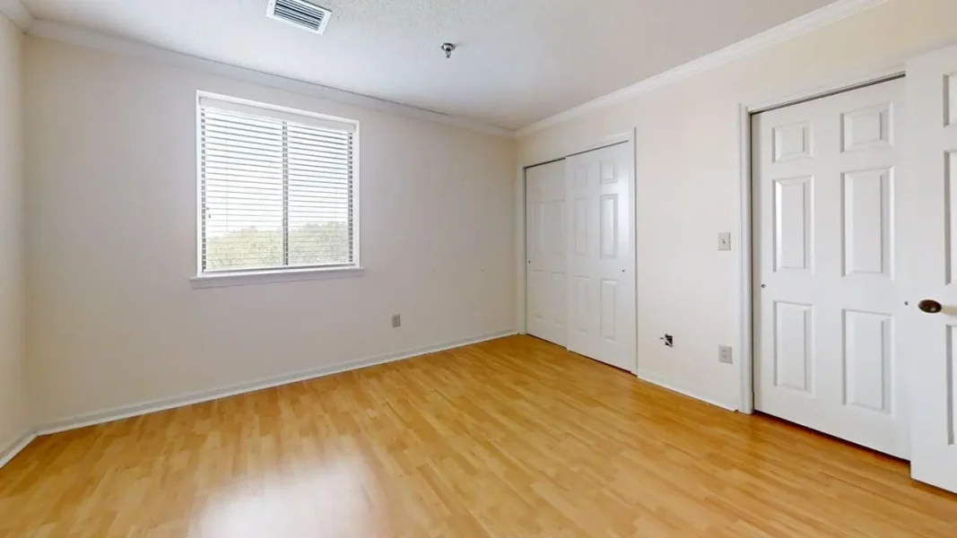 Empty bedroom with light wood floors, beige walls, a window with blinds, and two white closet doors along one wall.