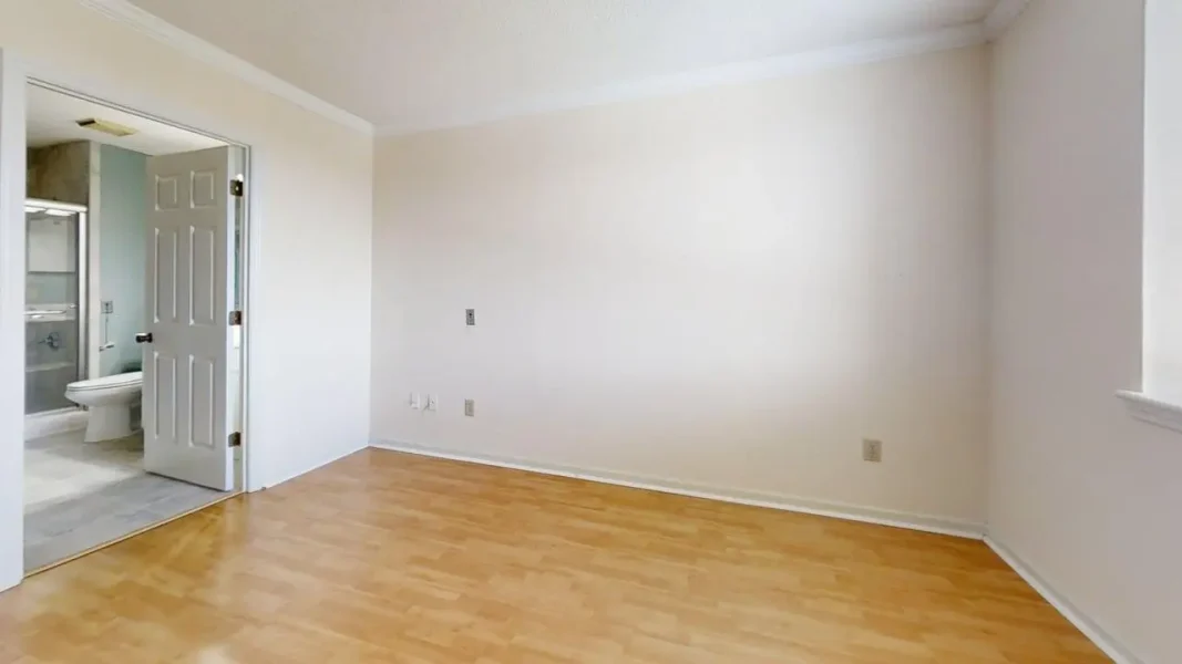 Empty room with light wood flooring and off-white walls; open doorway reveals a bathroom with a toilet and shower.