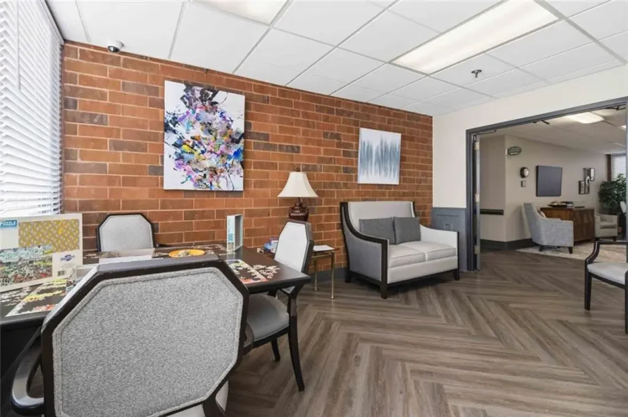 A waiting room with a brick accent wall, two abstract paintings, and a mix of gray upholstered chairs and a white sofa on a herringbone wood floor.