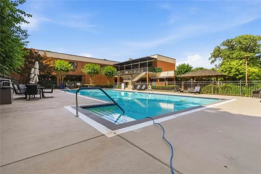 Outdoor swimming pool with a metal handrail, lounge chairs, and umbrellas, set beside a brick building and trees on a sunny day.