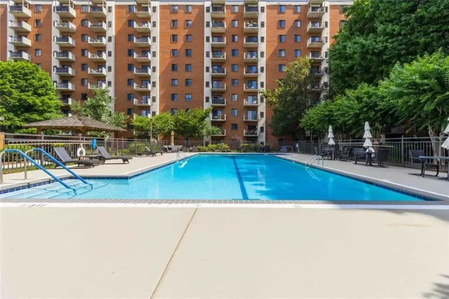 Outdoor communal pool area at a residential complex, with lounge chairs, umbrellas, and a brick apartment building in the background