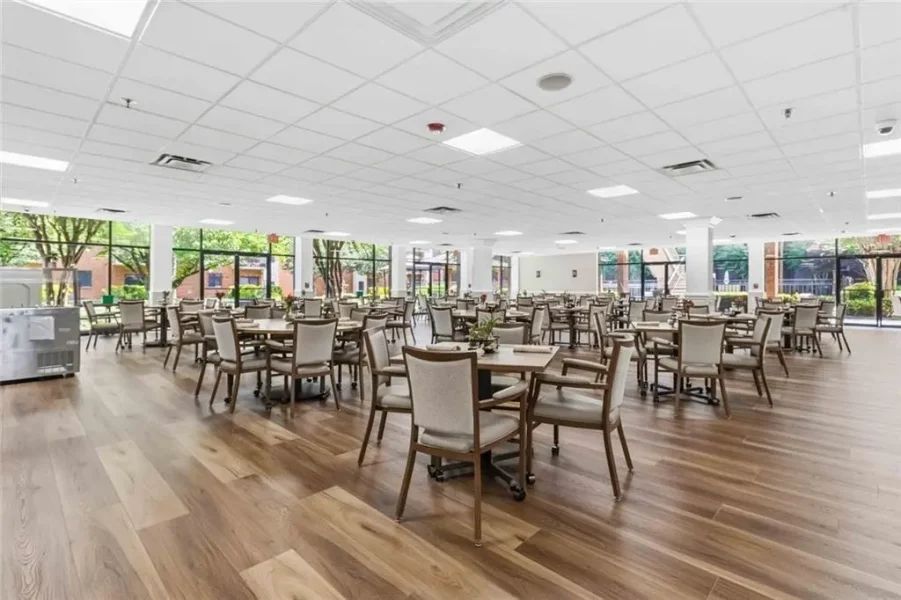 Bright cafeteria with many square tables and beige chairs on a wood floor, bathed in natural light from large windows.
