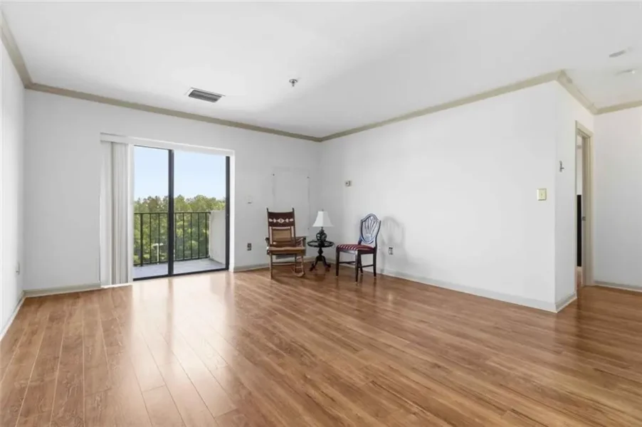 Spacious living room with light wood flooring, white walls, and a sliding glass door to a balcony; a small seating area with two chairs and a lamp on a side table in the corner.