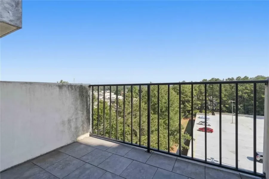 Balcony with gray tile floor and black metal railing overlooking a parking lot and tree line under a clear blue sky.