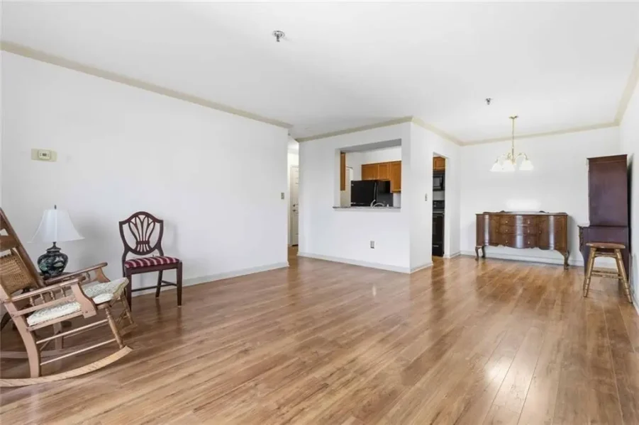 Spacious living room with hardwood floors, minimal furniture, rocking chairs, a wooden chair, and a vintage buffet against the wall; open kitchen pass-through visible in the center-right.
