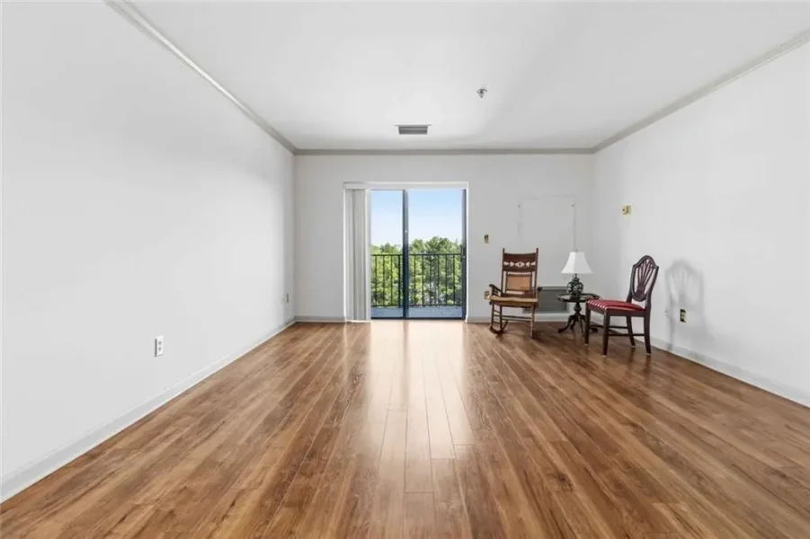 Empty living room with hardwood floors and white walls, sliding door to balcony with a view of trees outside.
