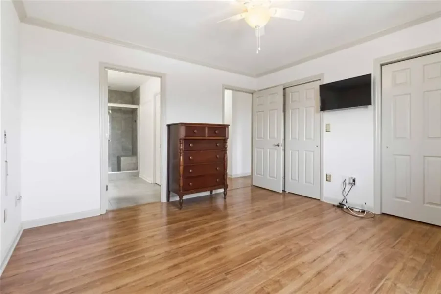 Empty bedroom with light hardwood floors, white walls, and a ceiling fan; a dark wooden dresser sits near the center, with multiple doors (closets and a bathroom) visible and a wall-mounted TV on the right.