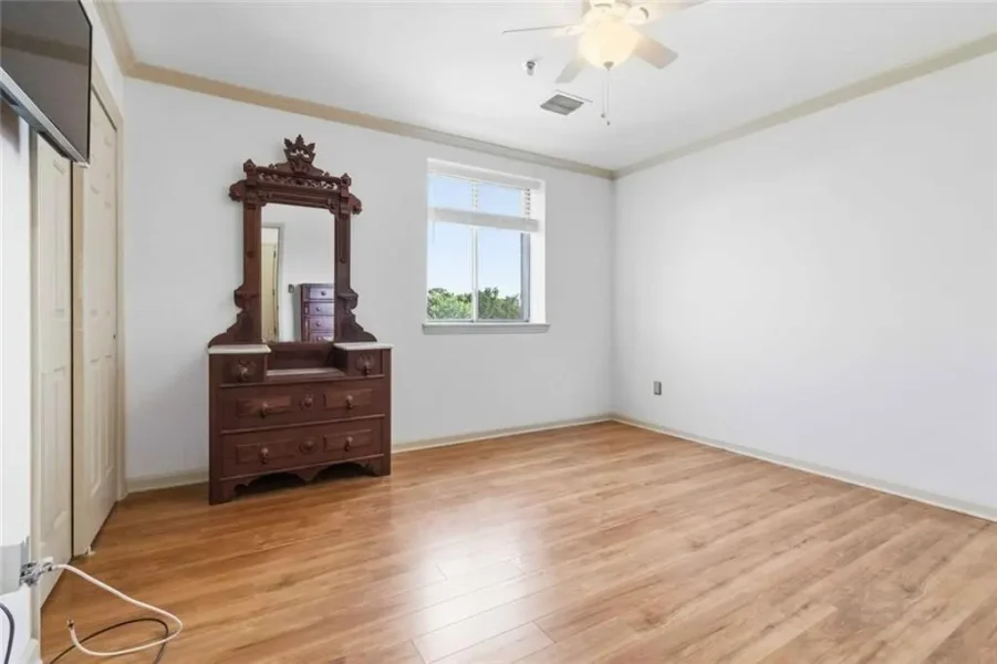 Empty bedroom with hardwood floors, white walls, and a dark wooden dresser with an ornate mirror near a window, plus a ceiling fan overhead.
