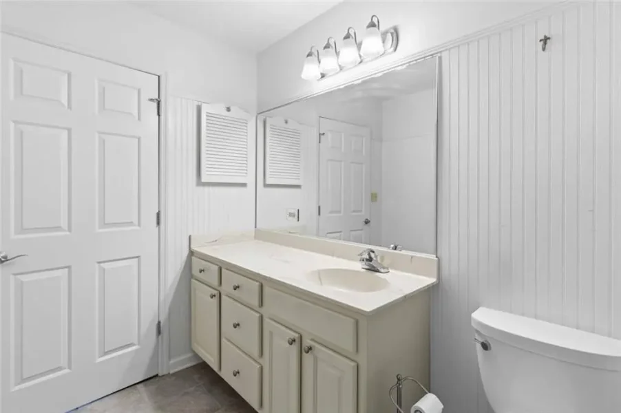 White bathroom with a long vanity, sink, and large mirror above; beadboard walls and a bright light fixture.