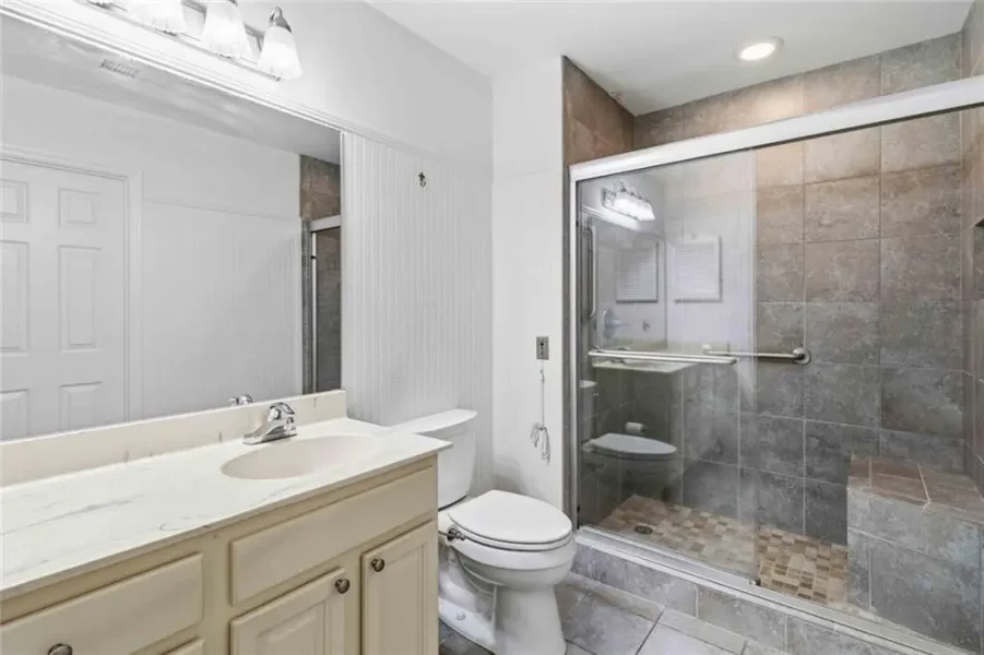 Bathroom with a long vanity and sink, mirror, and toilet beside a glass-enclosed walk-in shower with tiled walls and a built-in bench