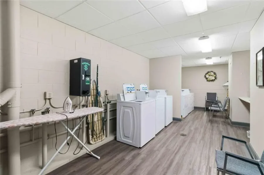 Laundromat-style utility room with multiple top-load washers and dryers, an ironing board, and folding area along a beige wall path to a seating nook at the back