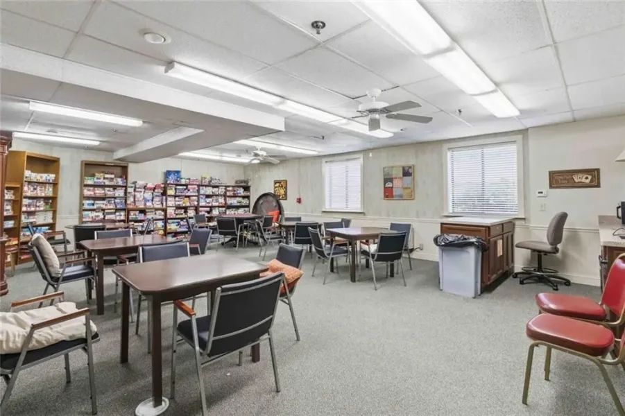 Community room with several square tables and chairs, bookshelves along the back wall, and a desk by a trash can under windows.
