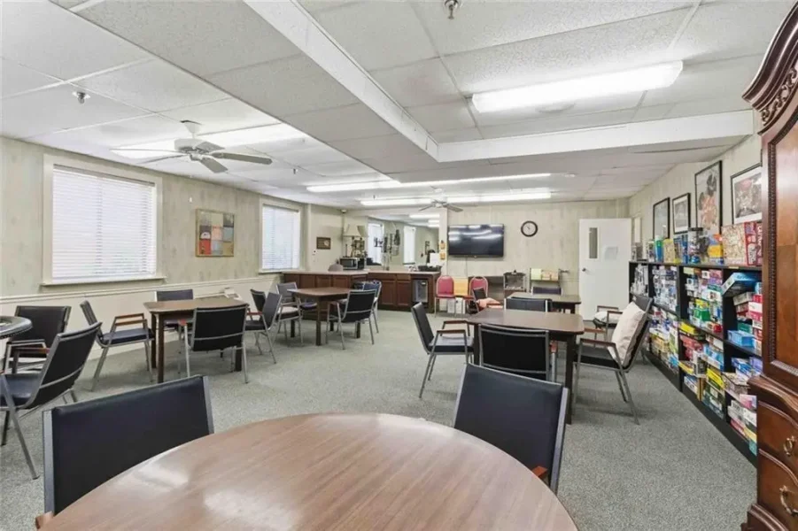 Community room with round and rectangular tables, chairs, and a game shelf lining the right wall; TV on the back wall and ceiling fans overhead.