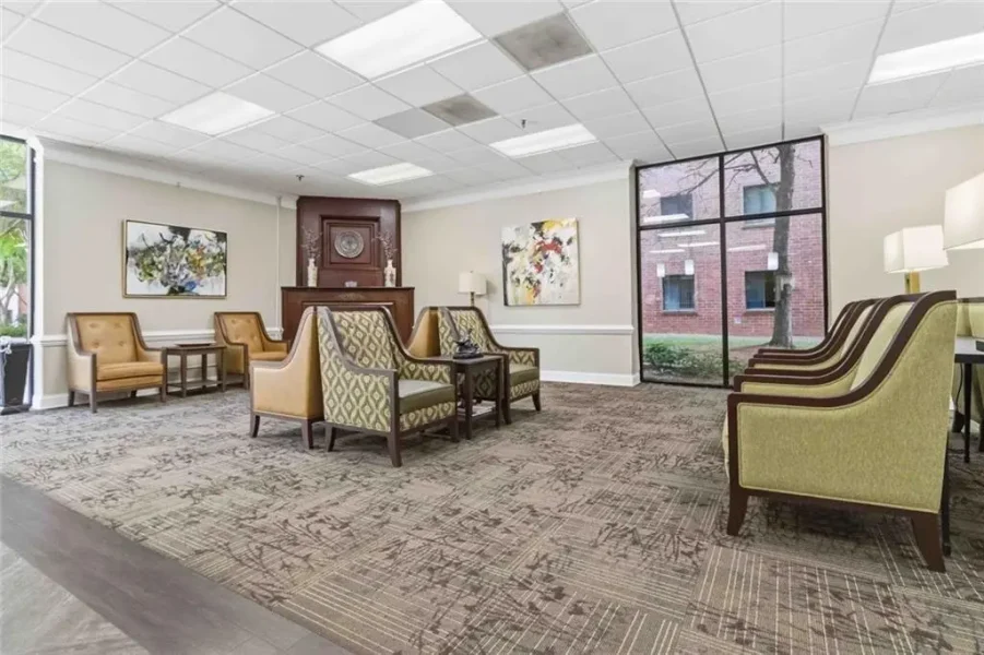 Lounge seating area with beige and patterned chairs around small tables, abstract art on the walls, and a wooden cabinet in the center.