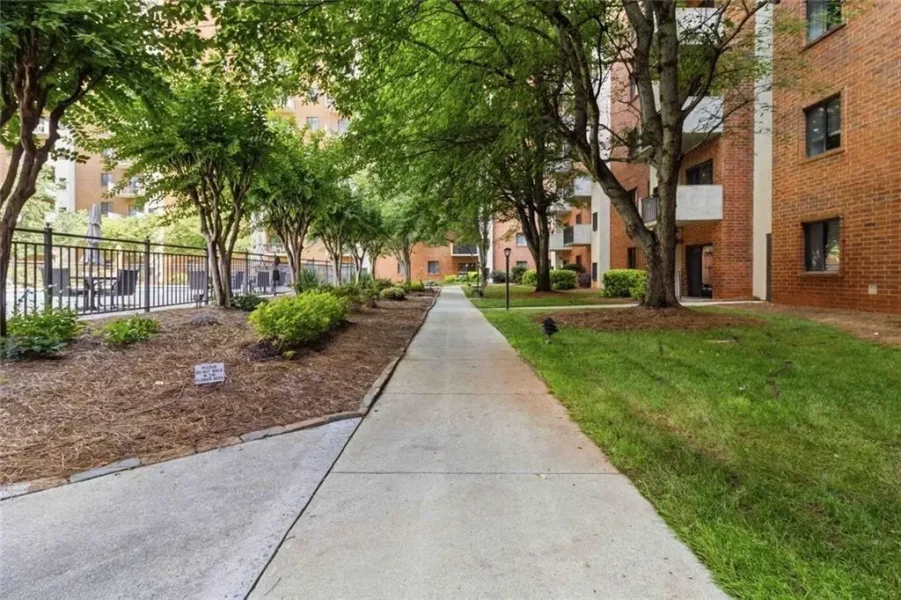 Concrete sidewalk cuts through a residential courtyard flanked by trees, shrubs, and brick apartment buildings on a sunny day, with a left seating area visible.