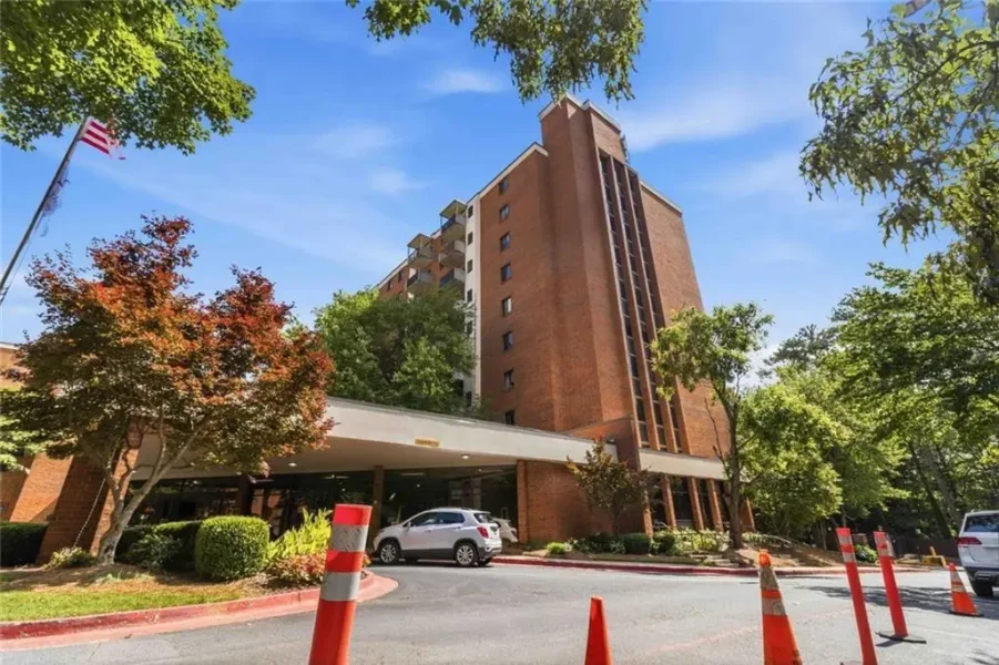 Tall brick hotel/apartment tower with a covered entrance, cars in the driveway, and orange traffic cones under a sunny sky.