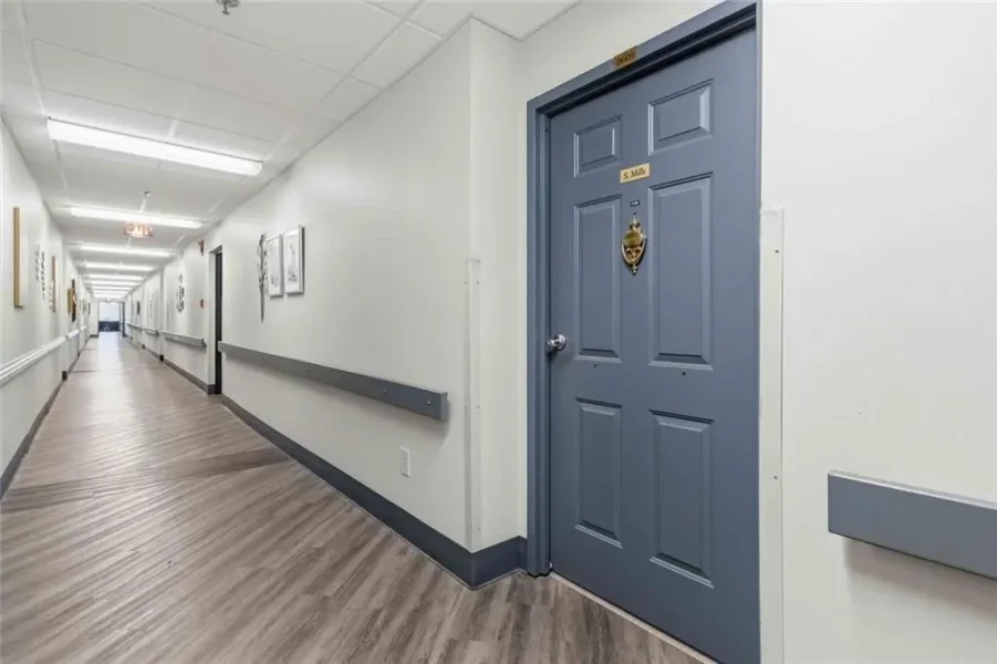 Long, brightly lit hospital hallway with pale walls, wood-toned flooring, and gray handrails. A blue door with a nameplate and door knocker is on the right.
