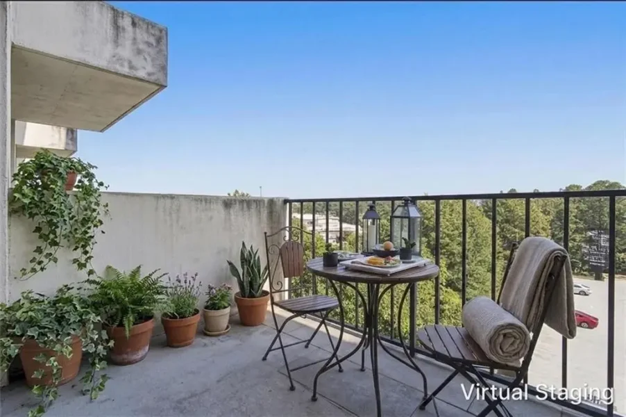 Cozy apartment balcony with potted plants along a concrete railing, a round metal table set for breakfast with two chairs and a rolled blanket draped over one chair, clear blue sky overhead.