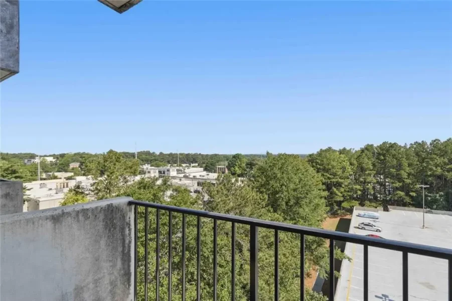 Balcony railing in the foreground with a treed, suburban landscape and low-rise buildings under a clear blue sky.
