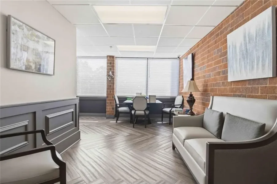 A waiting area with gray upholstered sofa and armchairs along a brick accent wall, abstract art, and a table lamp.”] ,