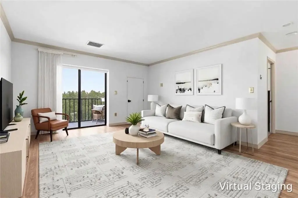 Bright living room with white sofa, two pillows, round coffee table, and a light rug; sliding door to balcony with outdoor chairs visible outside. (Virtual staging)